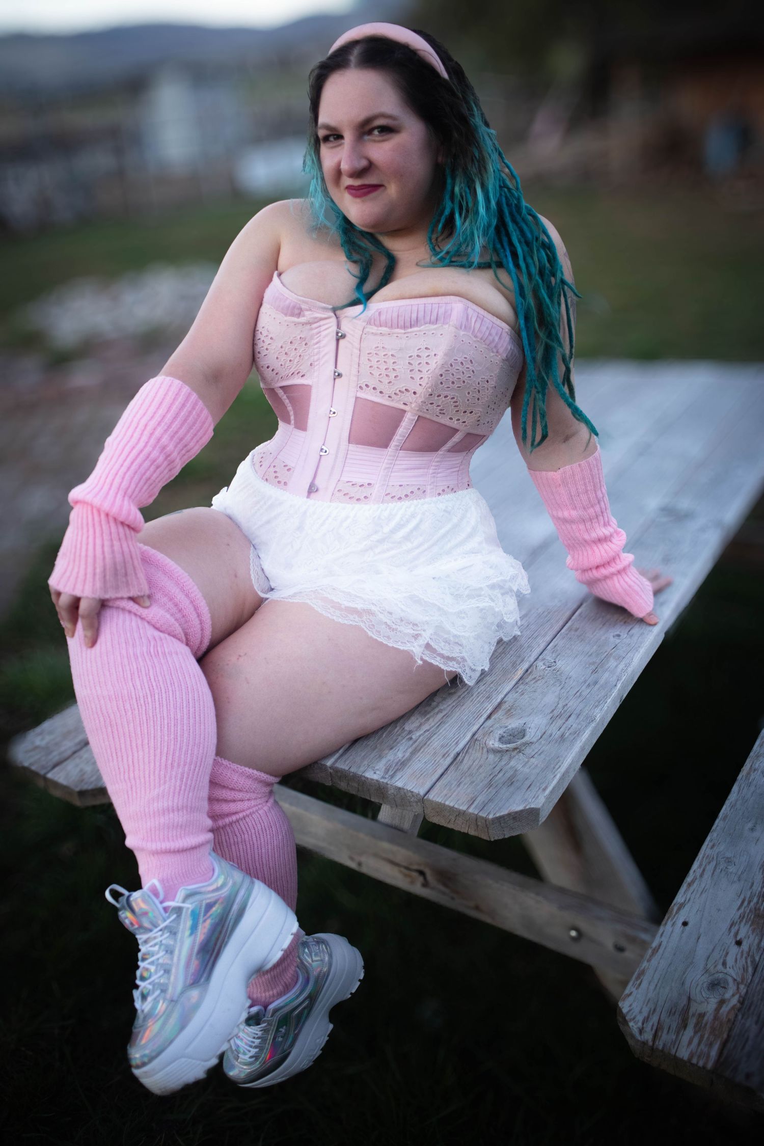 a photo of a person wearing a pink corset and white shorts with blue hair sitting on a picnic table seductively