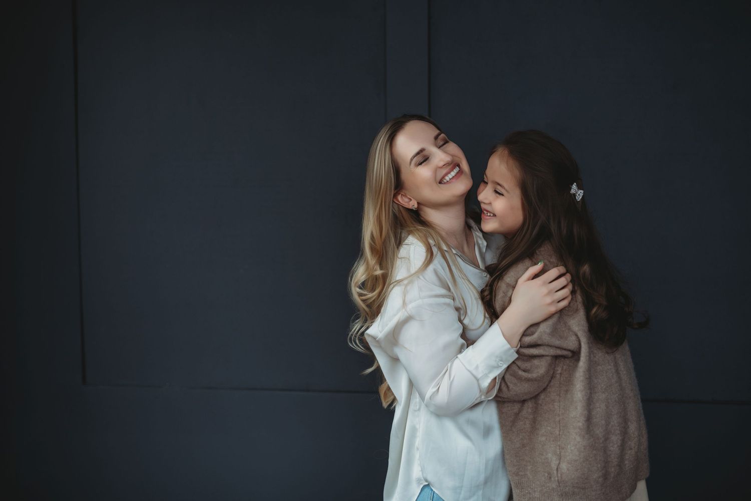 Two women laughing and embracing against a dark wall, wearing white and brown sweaters.