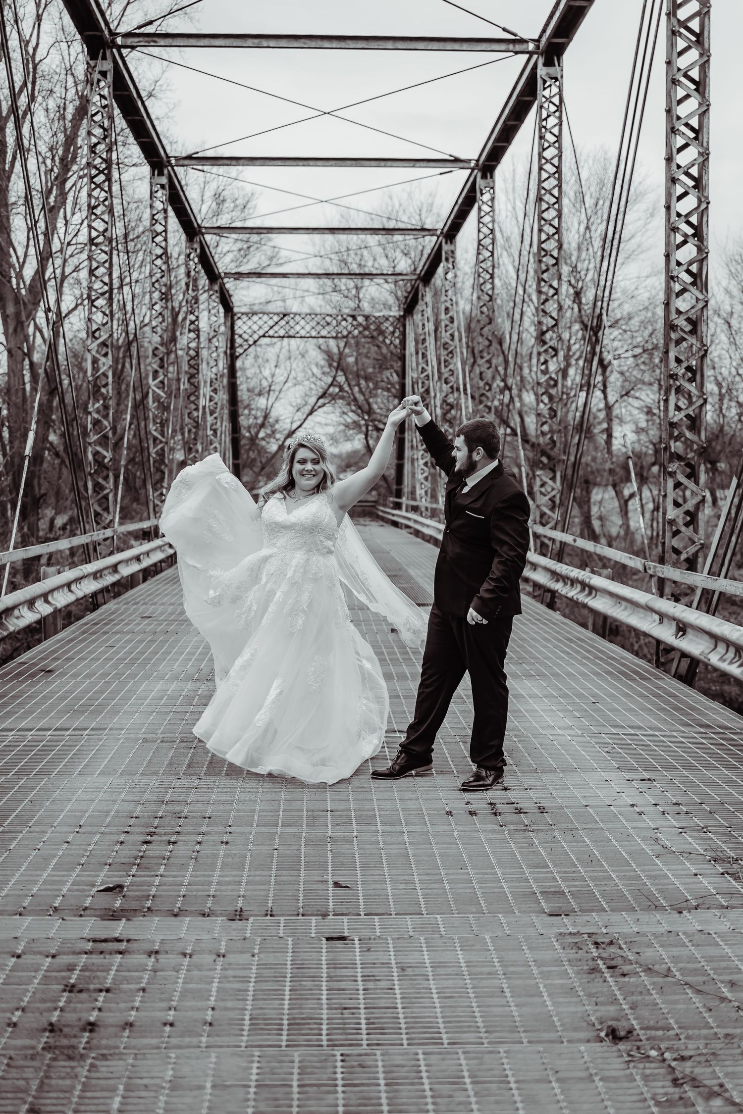A couple dancing on a metal bridge in black and white, with the bride twirling in a flowing white wedding gown.