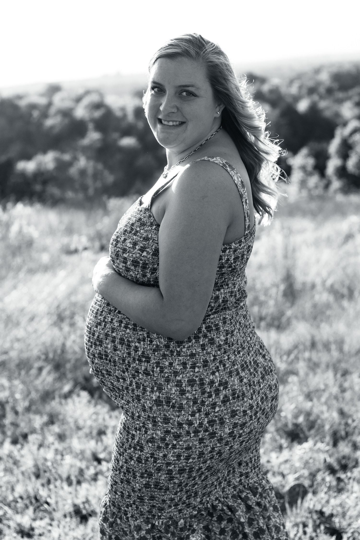 Black and white portrait of an expectant person posing in a floral sundress outdoors in a natural field setting.