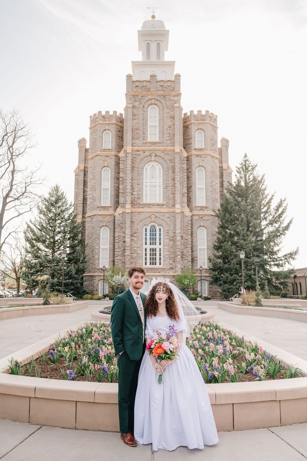 A couple poses in front of the Logan Utah Mormon Temple with spring tulips blooming in the foreground garden bed.
