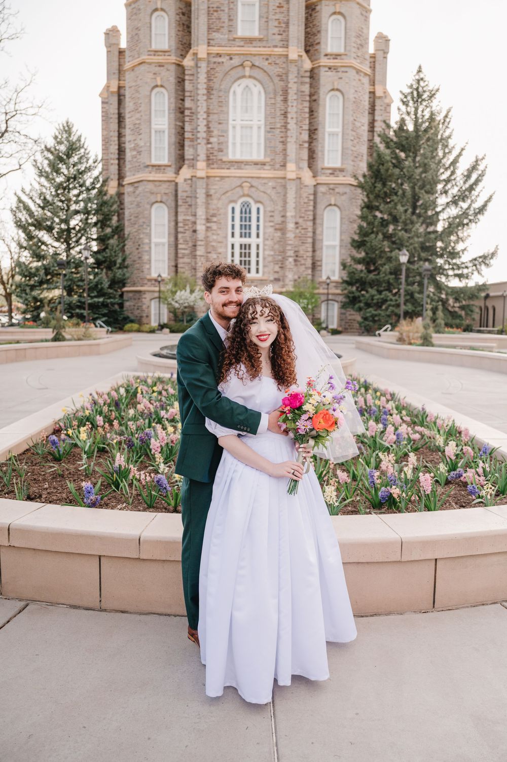 A couple in wedding attire share a joyful moment in front of a historic brick church surrounded by spring tulips.