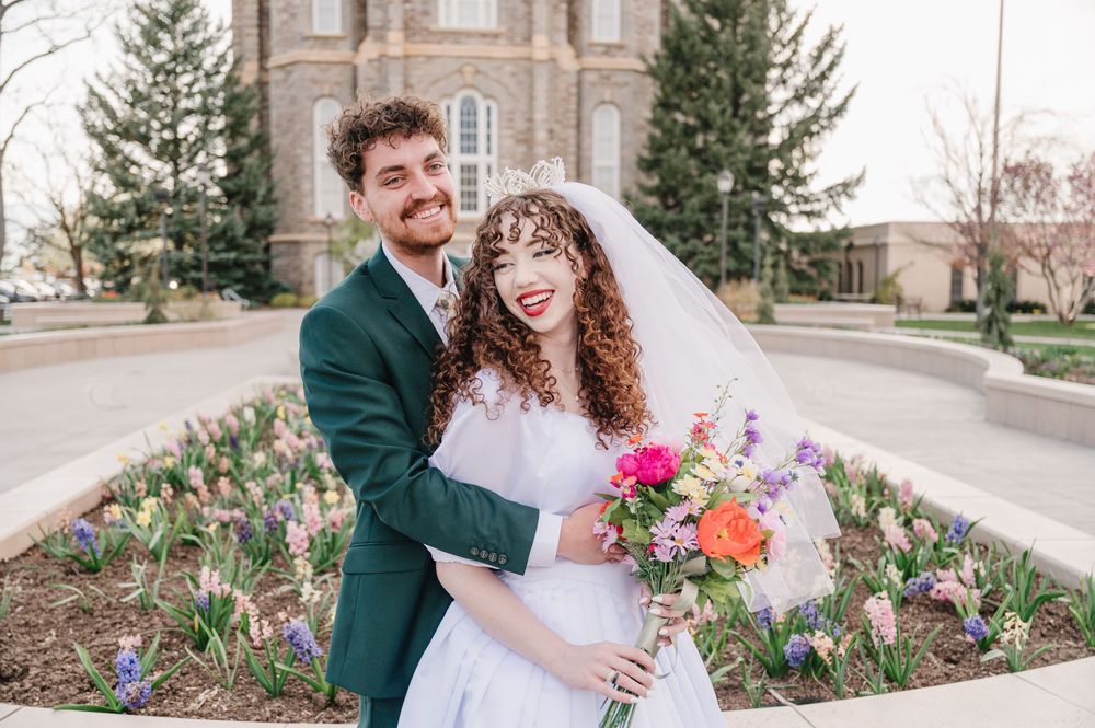A joyful couple shares a candid moment in front of a brick building surrounded by spring flowers in a garden.