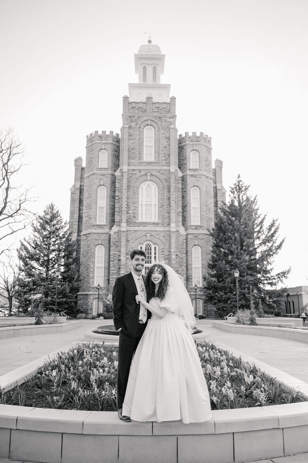 Black and white photo of a wedding couple standing in front of the historic Logan Utah Temple with blooming flowers.