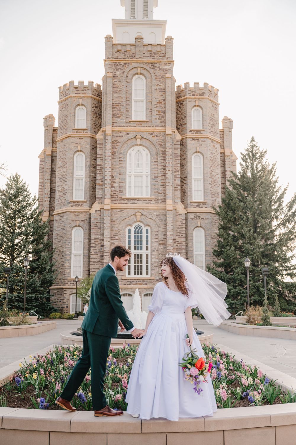 A couple in wedding attire walk hand in hand outside a grand stone temple with elegant grounds and spring flowers.