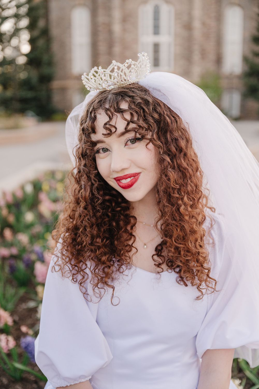 A bride wearing a white wedding dress with puff sleeves, delicate tiara and veil, showing curly brown hair and red lipstick.