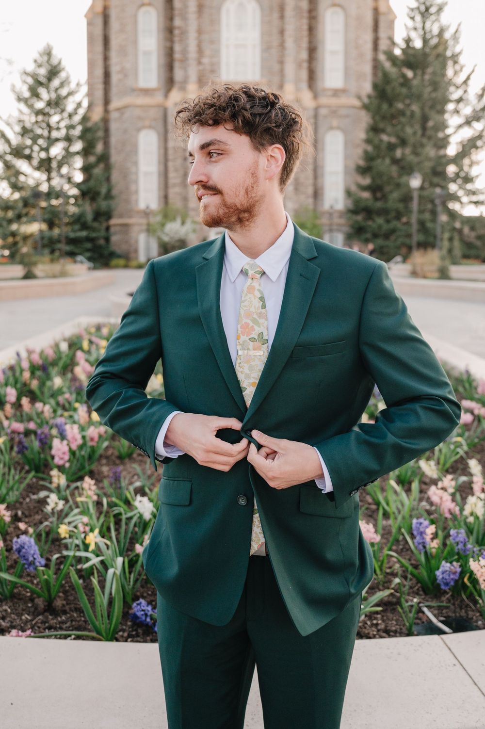 A person in an emerald green suit adjusts their jacket while standing in a garden with purple flowers.