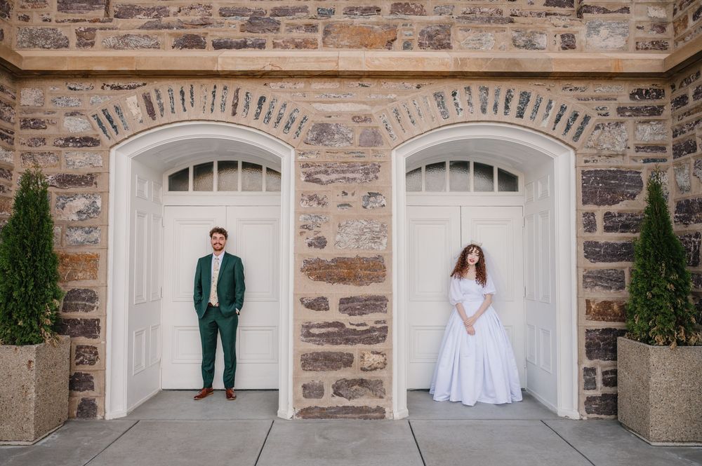Two figures stand in separate white arched doorways against a rustic stone wall with potted cypress trees on either side.