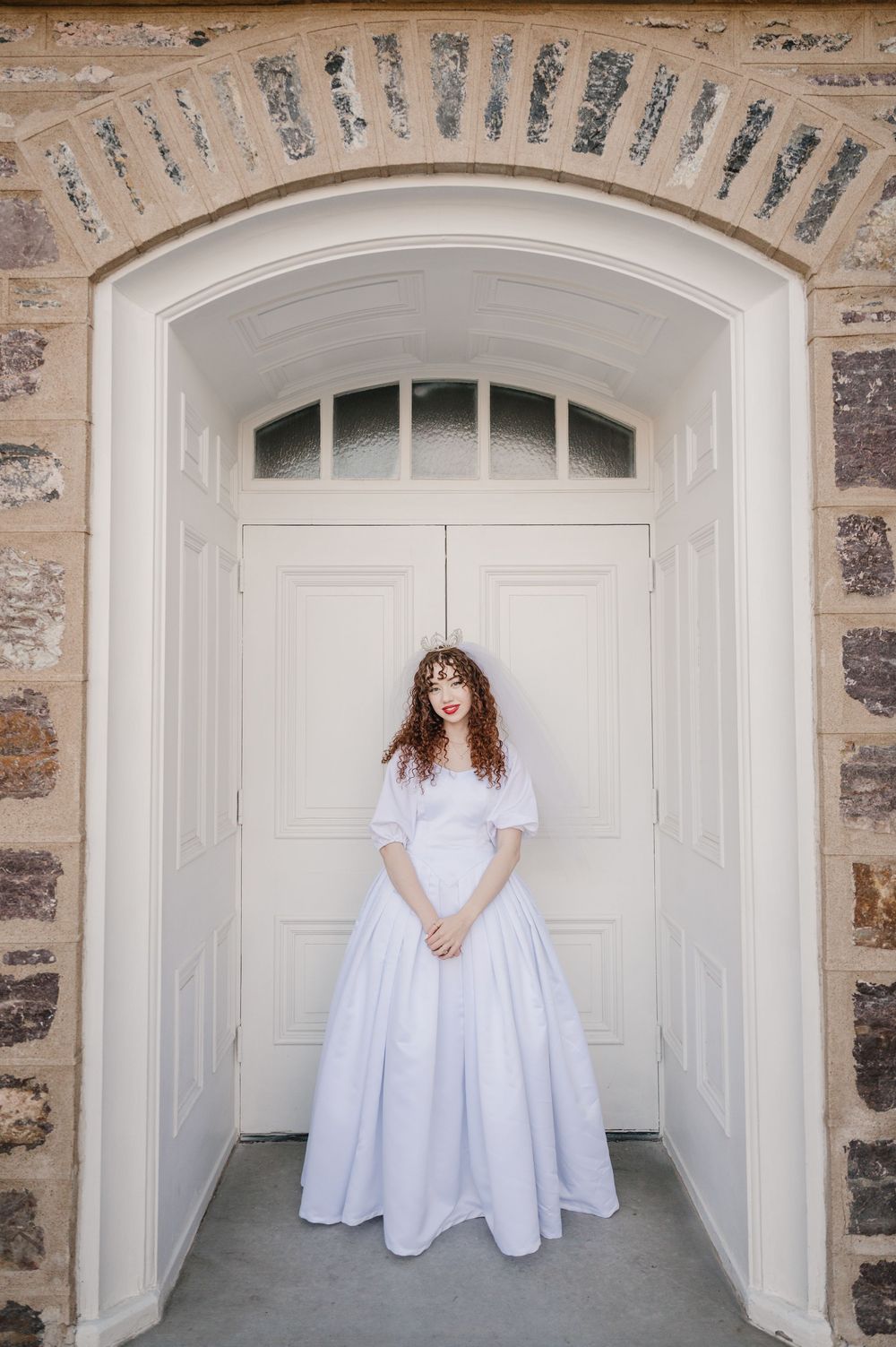 A person wearing a long white dress stands in an arched stone doorway with vintage architectural details.