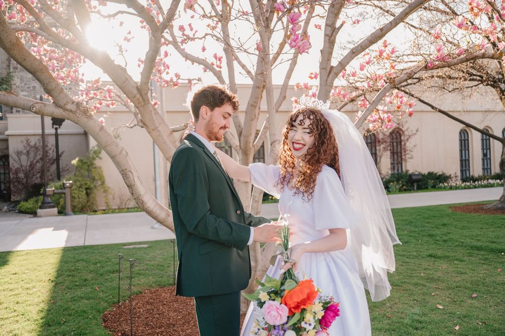 A newlywed couple stands beneath blooming cherry blossoms on their wedding day with colorful spring bouquet.