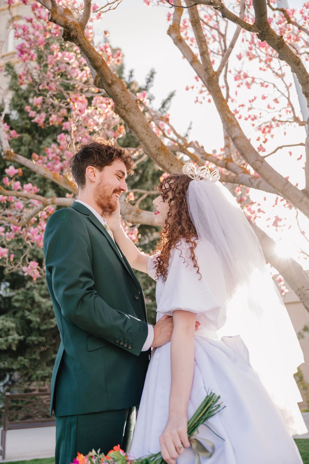 A springtime wedding portrait under blooming cherry blossoms features traditional white dress and emerald green suit.