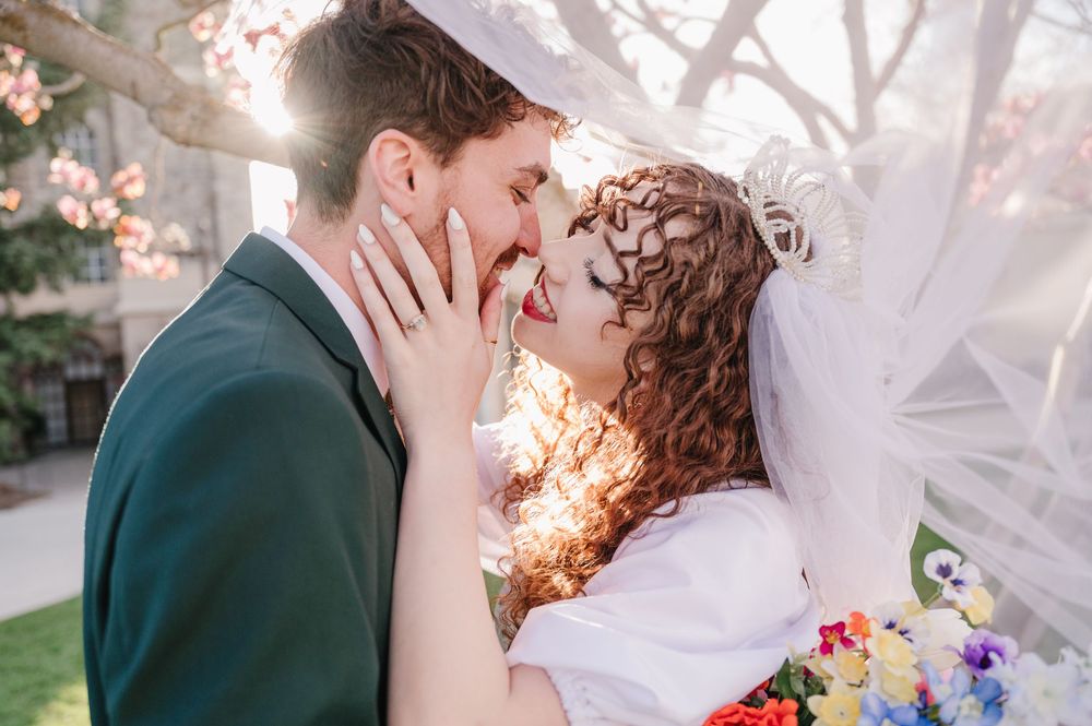 A couple in wedding attire share a tender moment outdoors with sunlight streaming through trees in the background.