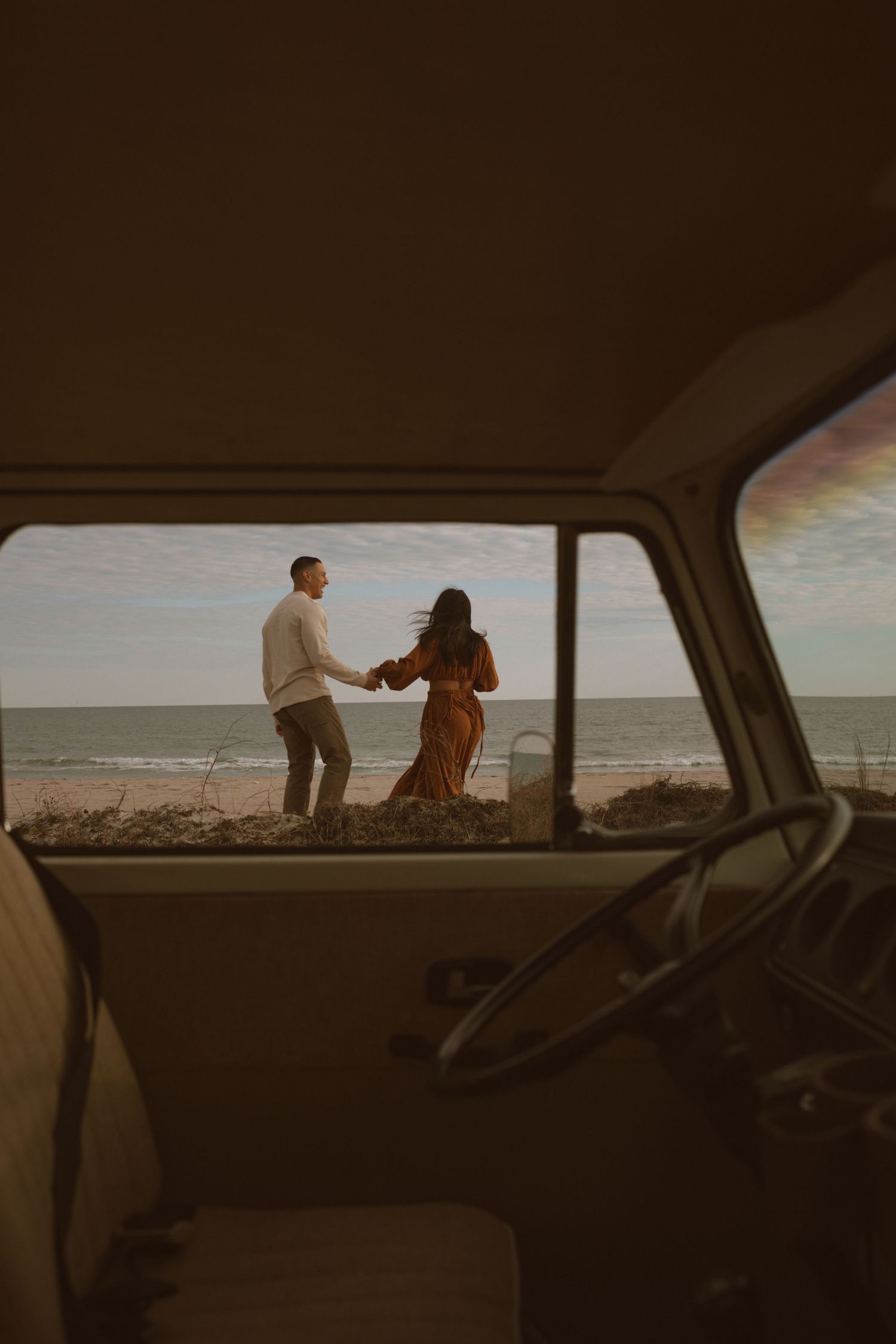 A scenic view through a car window shows figures silhouetted against a coastal landscape at sunset.