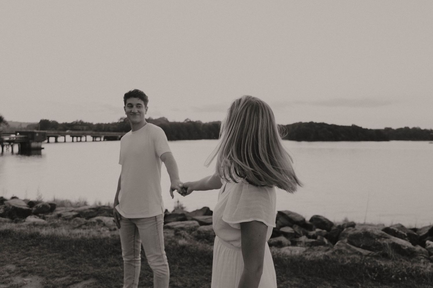 A couple holds hands while standing by a waterfront with rocks and a lake view at sunset.