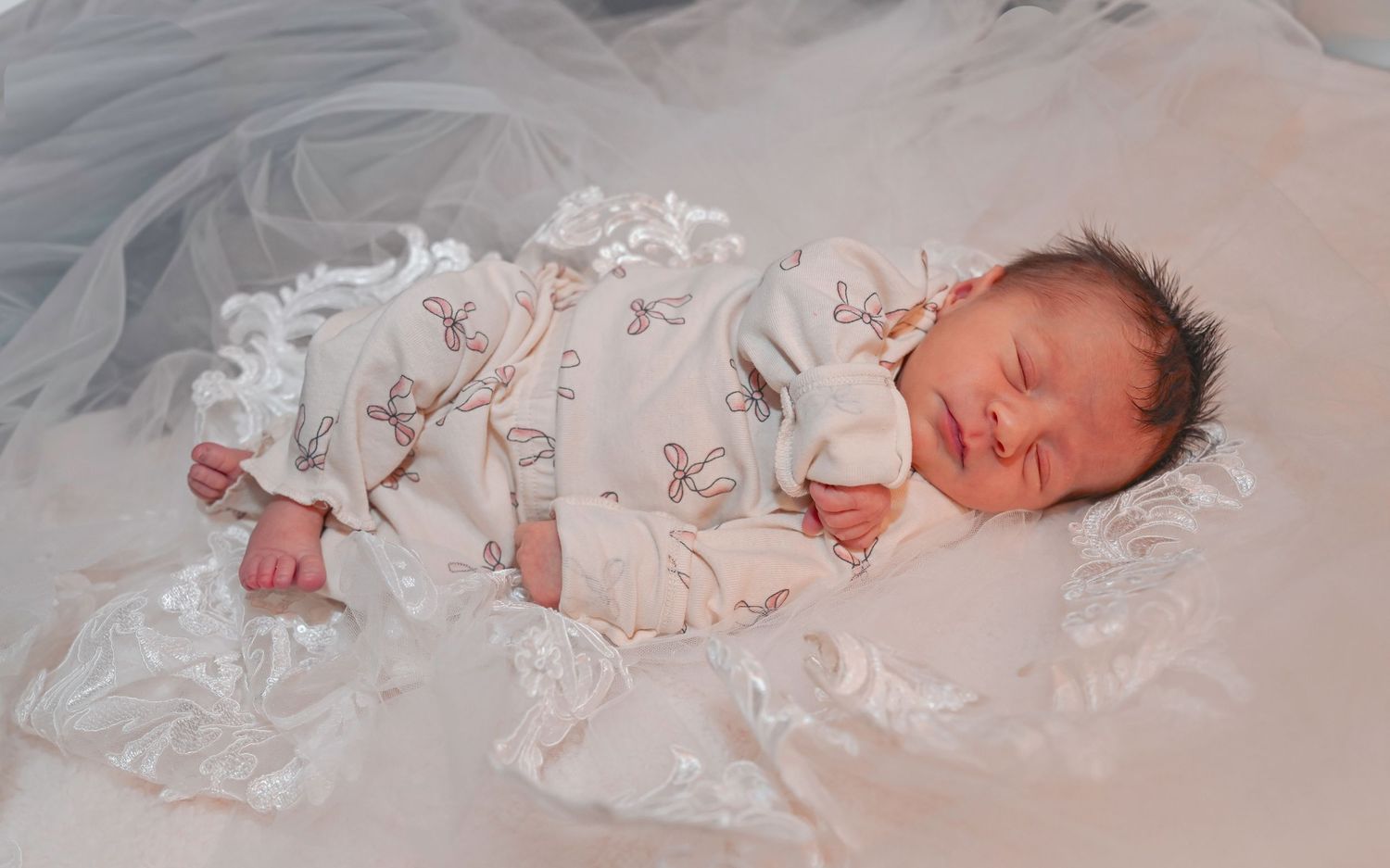Peaceful newborn baby sleeping in white lace outfit on soft white fabric with delicate details.