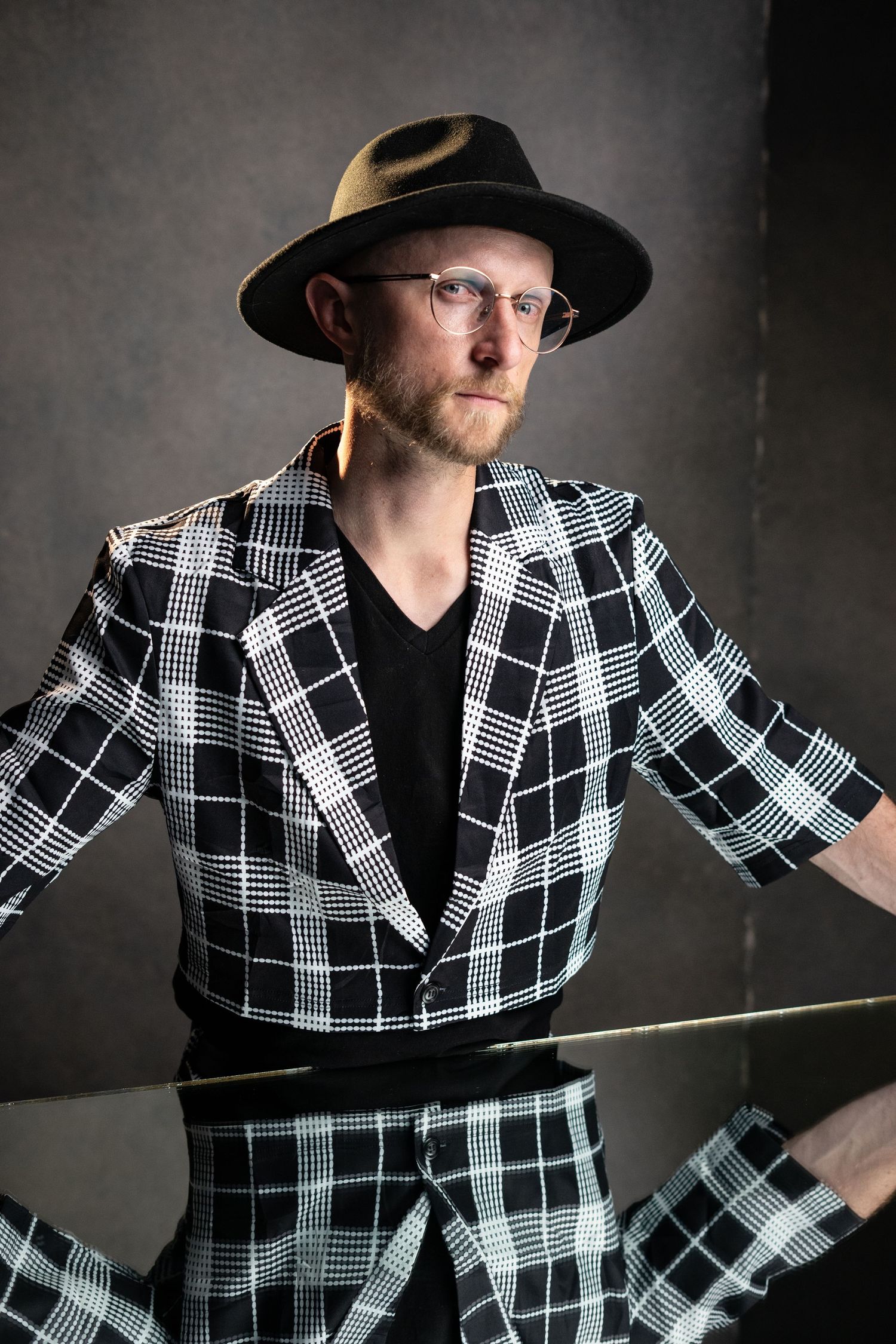 A performer in a black and white plaid suit and fedora hat strikes a dramatic pose in artistic studio lighting.