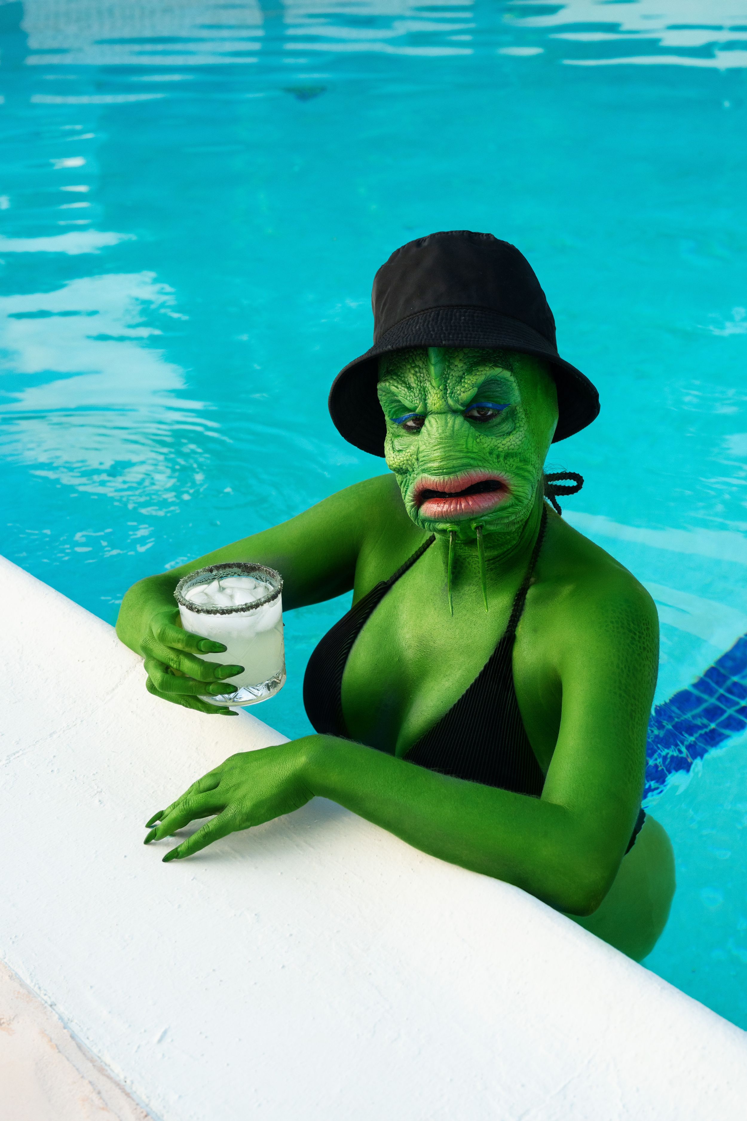 Green lizard monster sitting near the pool with a drink in hand and sunglasses enjoying the sun.