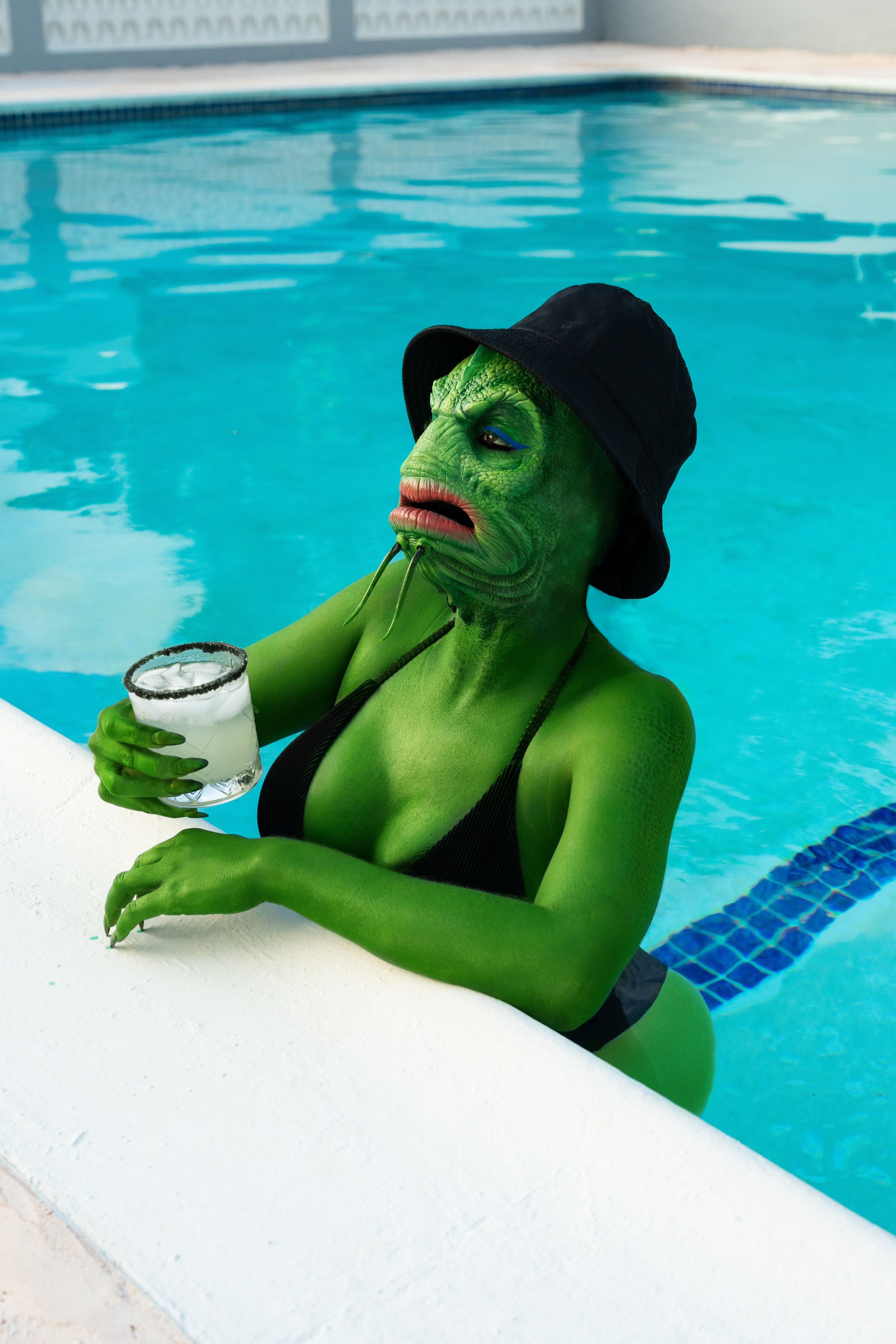 Green lizard monster sitting near the pool with a drink in hand and sunglasses enjoying the sun.