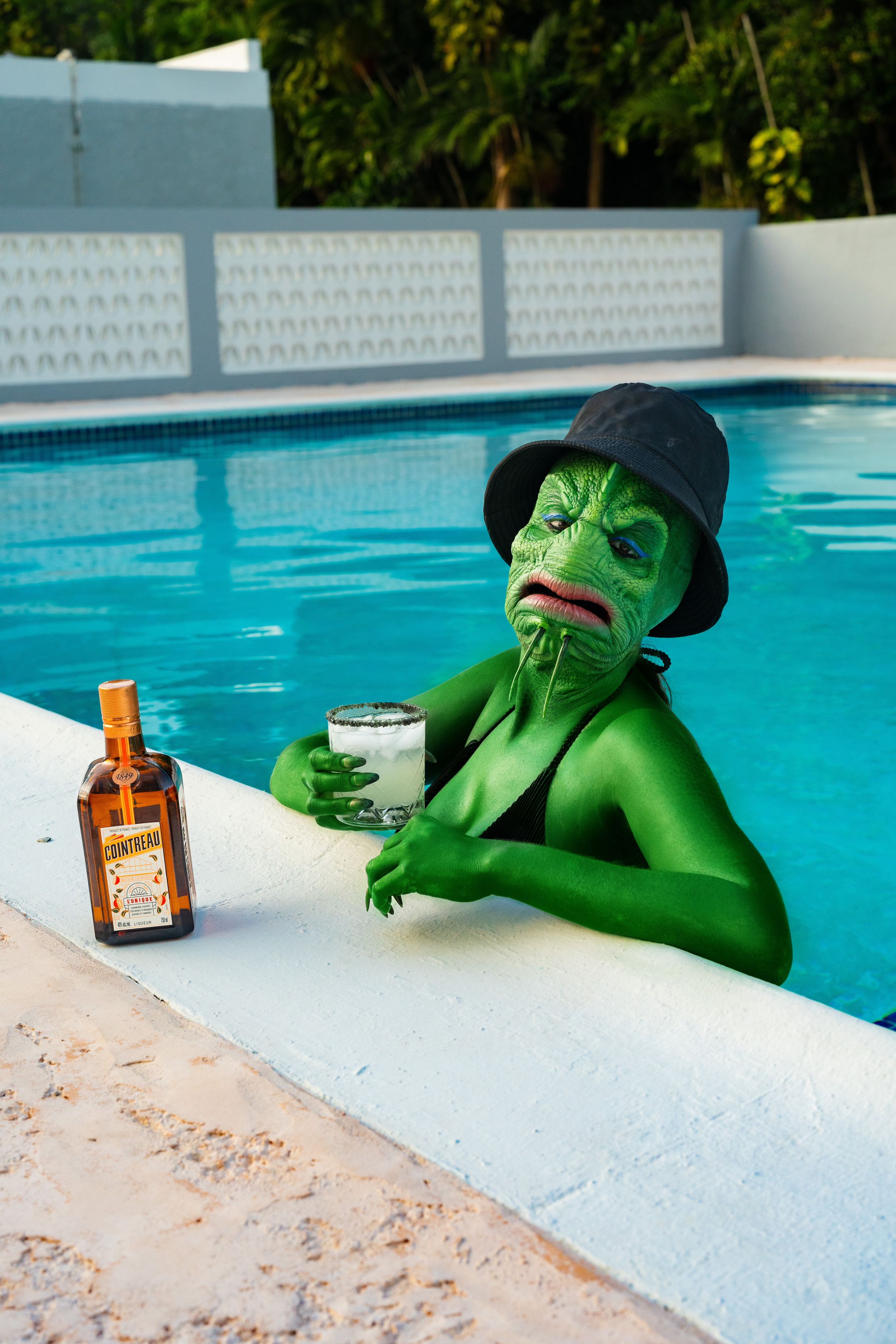 Green lizard monster sitting near the pool with a drink in hand and sunglasses enjoying the sun.