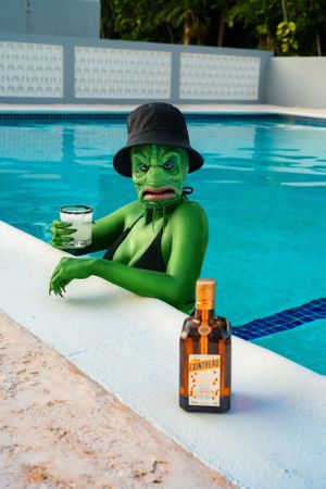 Green lizard monster sitting near the pool with a drink in hand and sunglasses enjoying the sun.