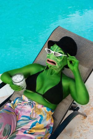 Green lizard monster sitting near the pool with a drink in hand and sunglasses enjoying the sun.