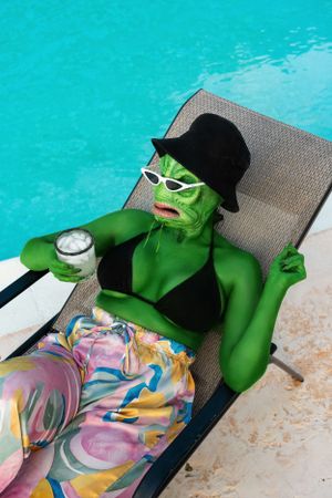 Green lizard monster sitting near the pool with a drink in hand and sunglasses enjoying the sun.