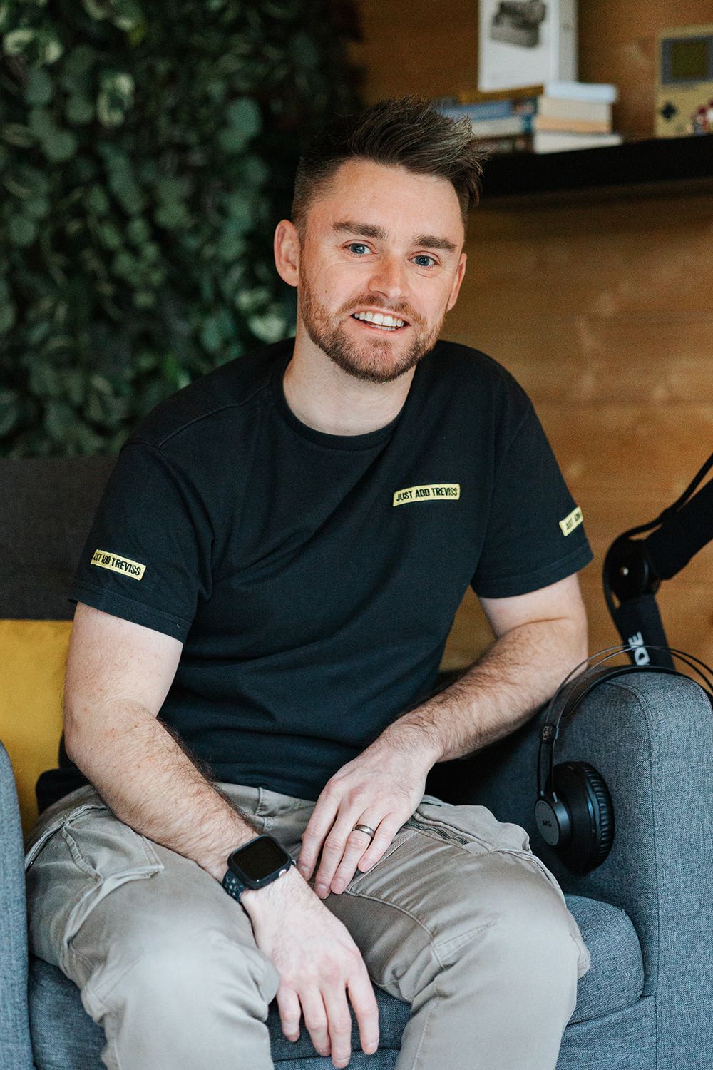 Casually dressed person in black t-shirt and gray pants sits relaxed in front of indoor greenery backdrop.