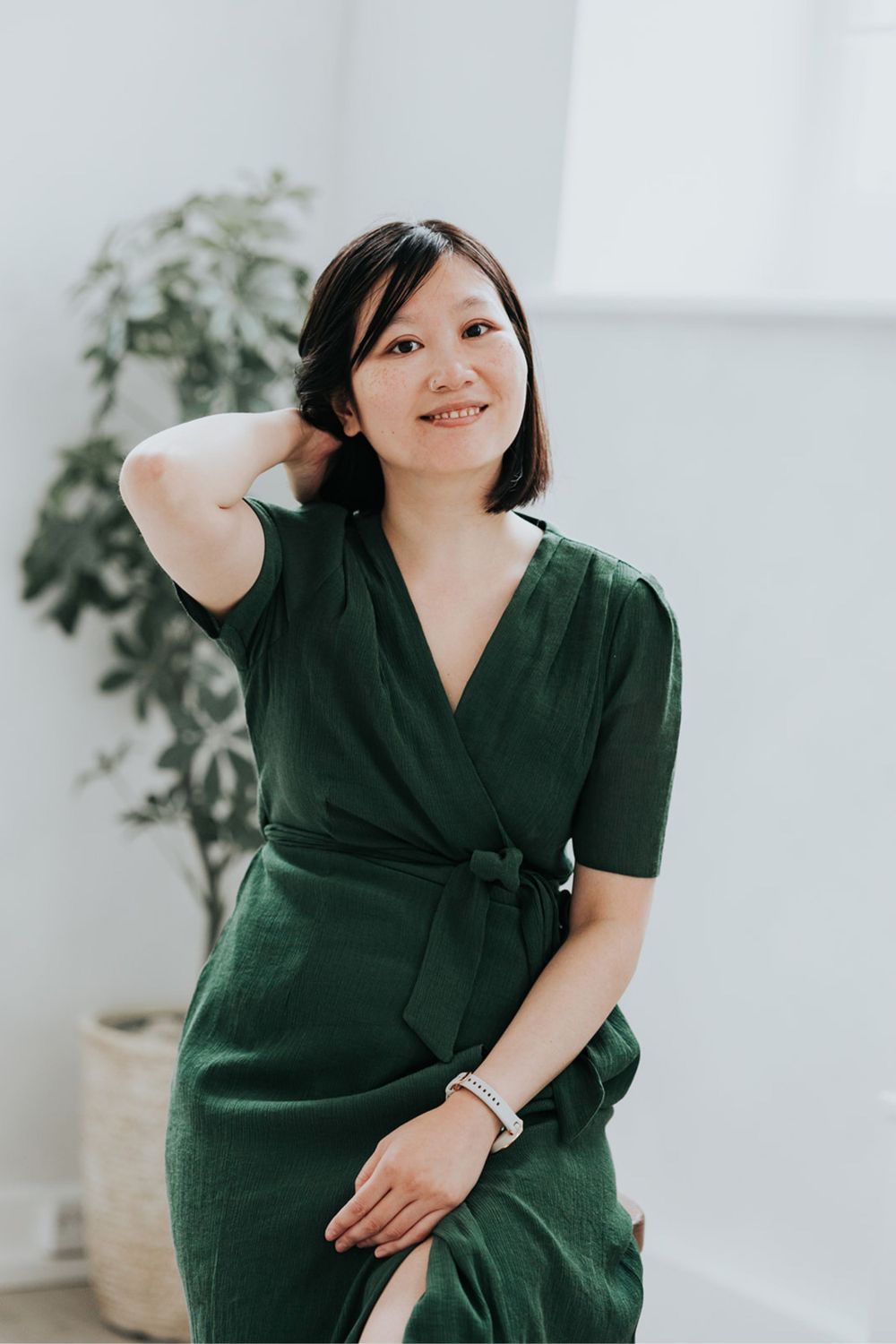 A person wearing a green wrap dress smiles while standing against a white wall with a potted plant in the background.