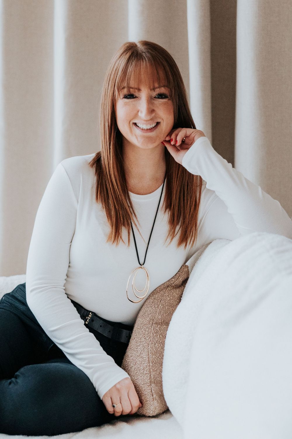 A person in a white long-sleeve top and black pants sits casually on a couch wearing a pendant necklace.