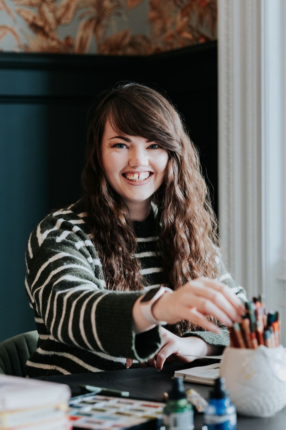 A creative workspace scene showing someone in a striped sweater seated at a desk with art supplies and materials.