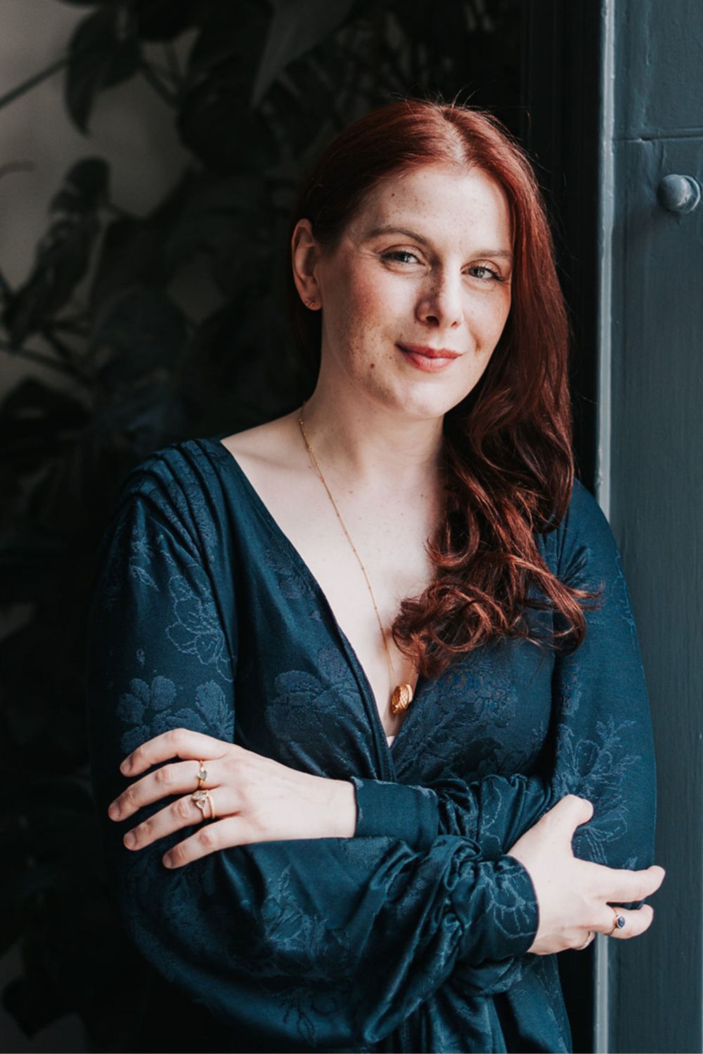Portrait of someone in a dark blue floral wrap dress with long red hair against a dark background.