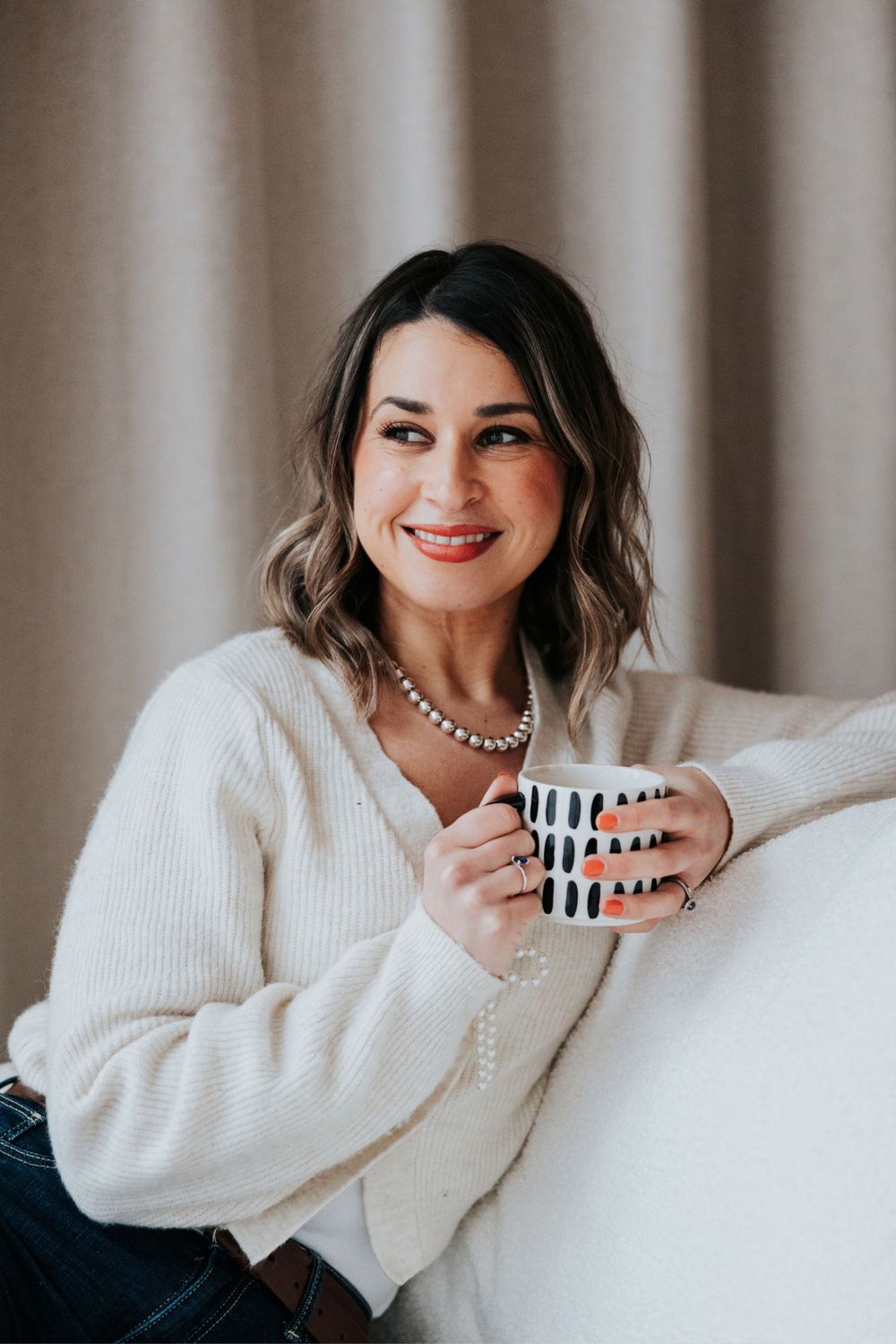 A smiling woman in a cozy white sweater holds a black and white patterned coffee mug while posing against a neutral backdrop.