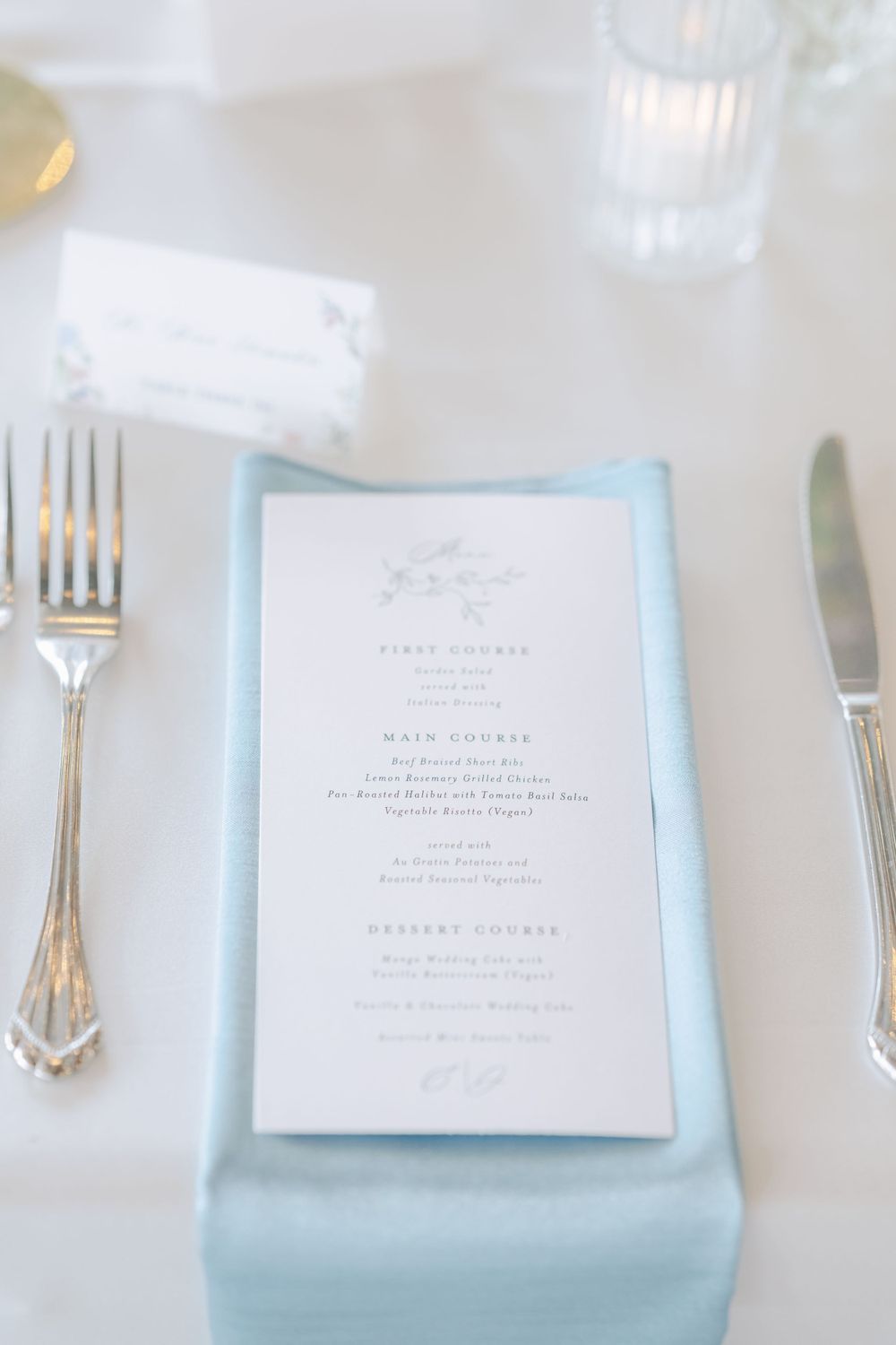 Elegant wedding dinner menu card placed on light blue napkin with silver cutlery on white tablecloth.
