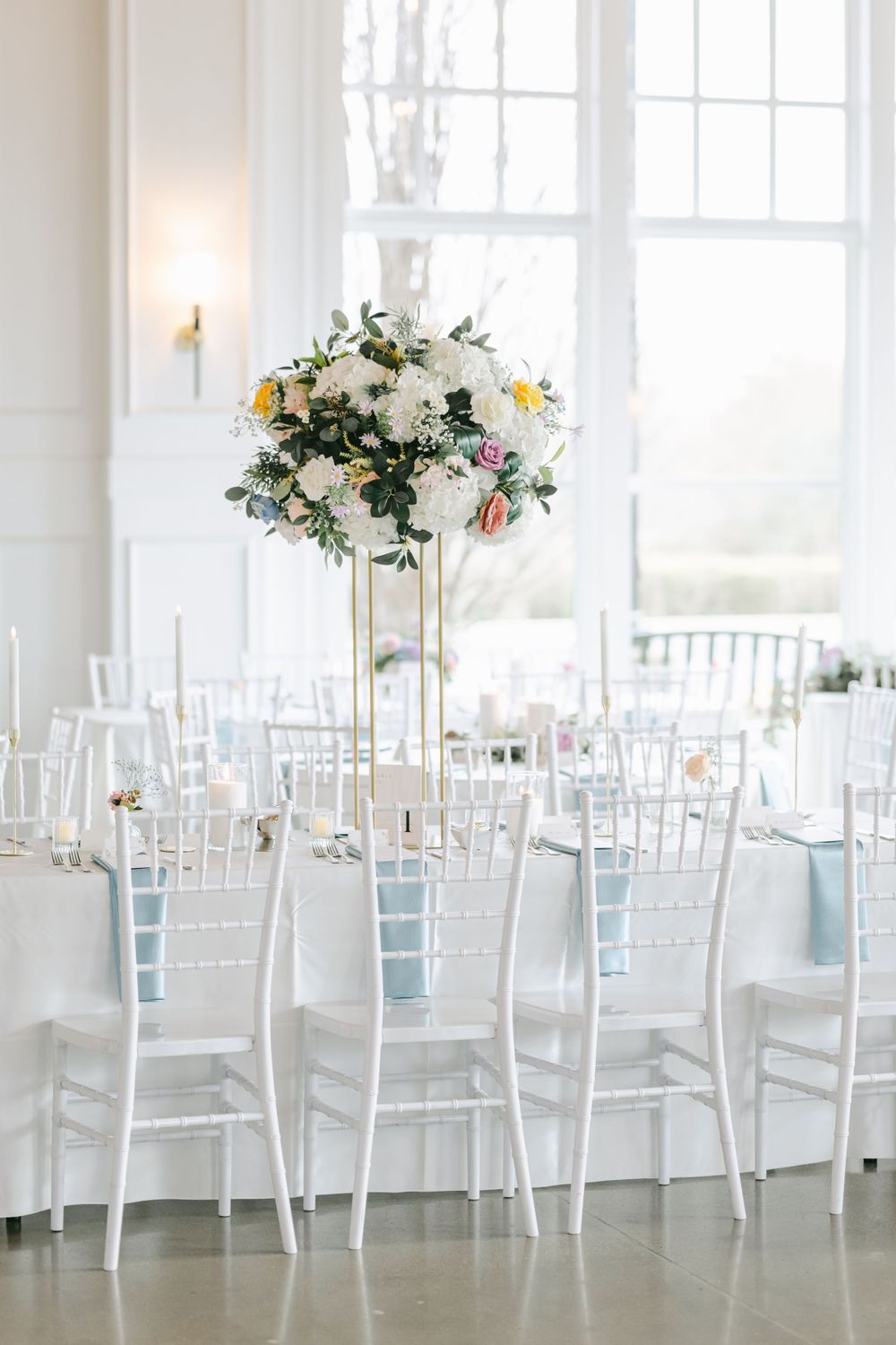 Elegant white wedding reception setup with tall floral centerpiece and white Chiavari chairs with light blue cushions.