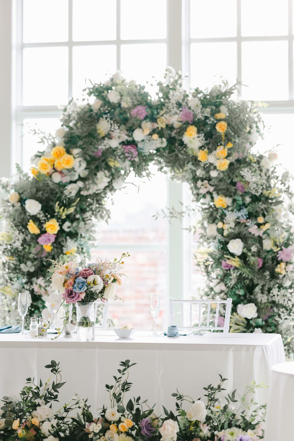 Elegant floral arch with white, yellow and pink roses decorates a wedding reception table against bright windows.