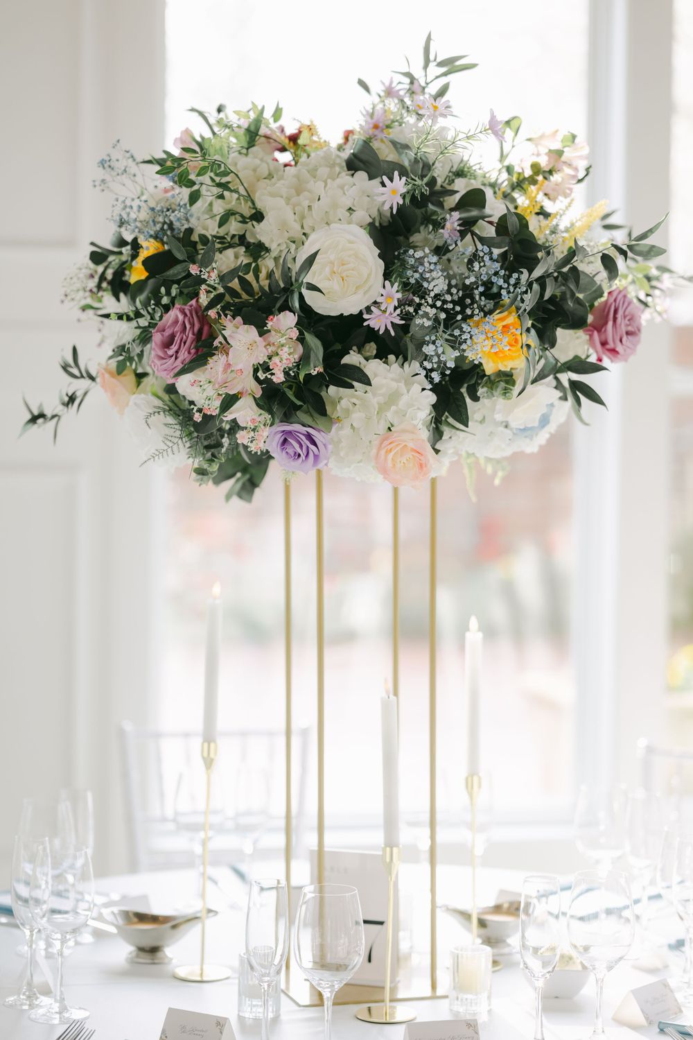 Elegant tall floral centerpiece with white roses, purple and yellow flowers on gold stands for wedding table decor.