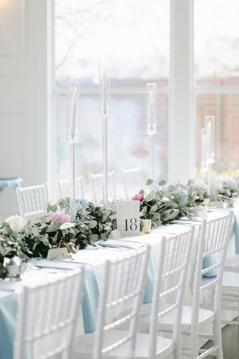 Elegant wedding table setting with white Chiavari chairs, light blue linens, tall glass candlesticks and eucalyptus garland.