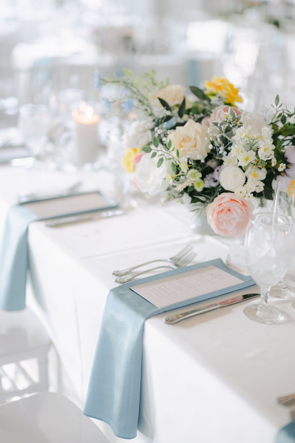 Elegant table setting with white and pink floral centerpiece, light blue napkins, and white linens for a wedding reception.