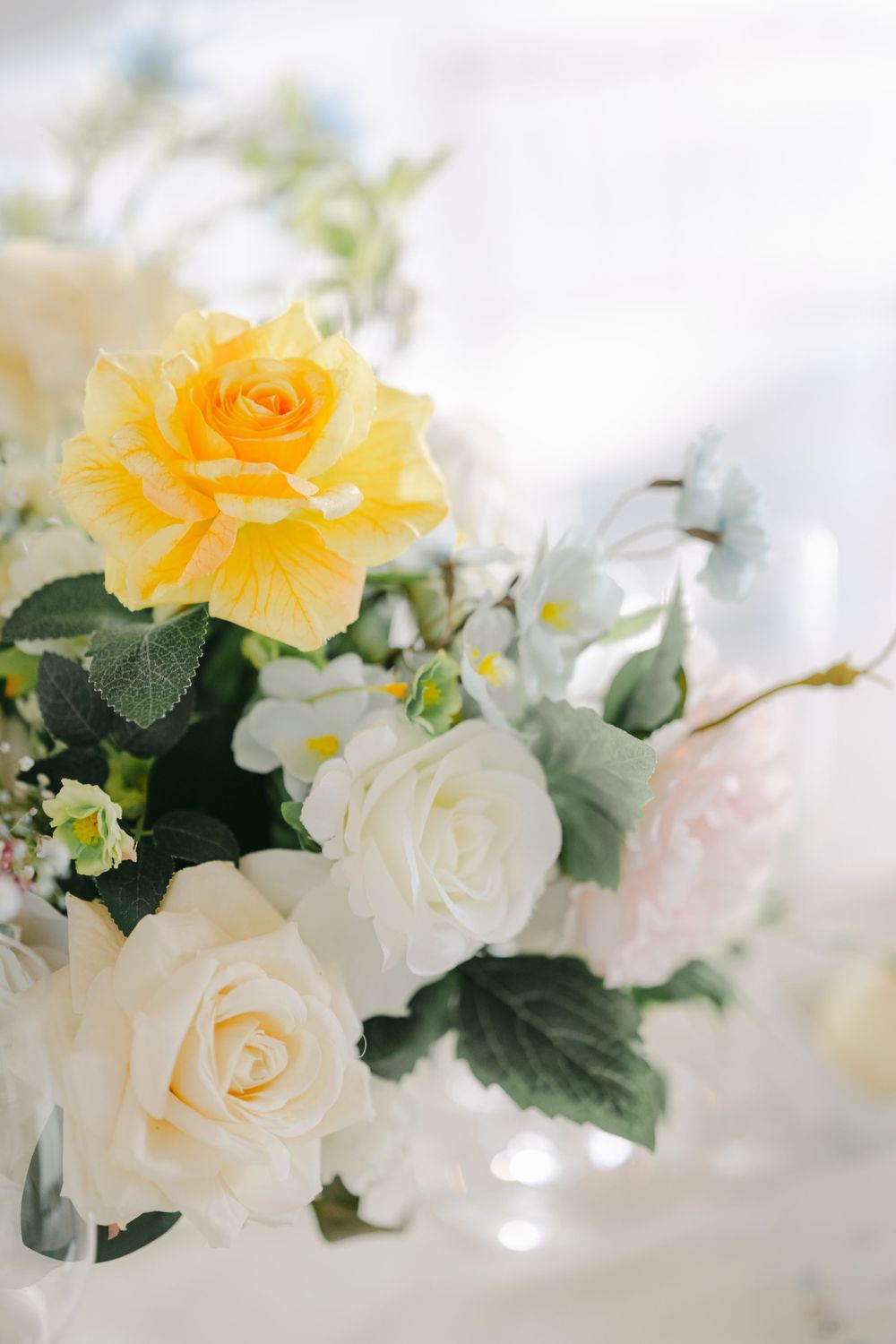 Delicate arrangement of yellow and white roses with green leaves in soft focus against a light background.