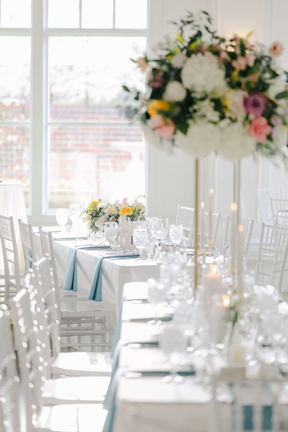 Light-filled wedding reception venue with white chiavari chairs, blue table runners and elevated floral arrangements.