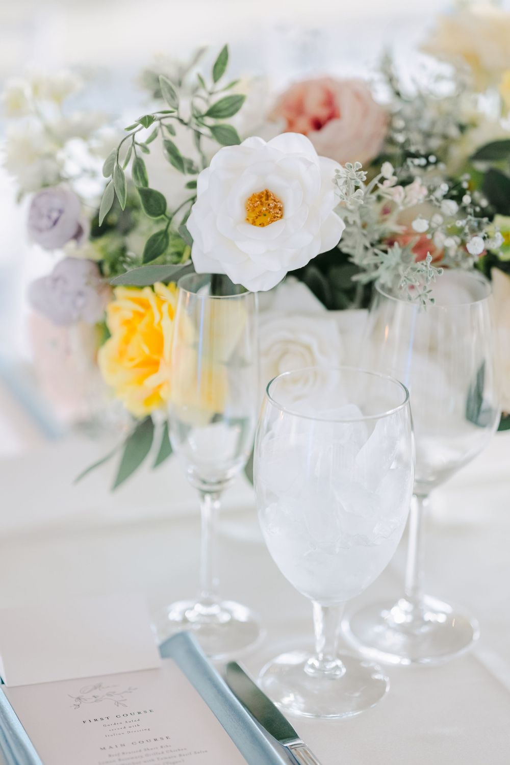 Delicate floral centerpiece with white and yellow blooms next to empty wine glasses on an elegant table setting.
