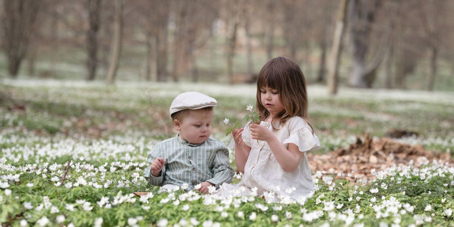 Ett ungt syskonpar sitter på en äng av vitsippor och tittar på blommorna. Syskonfotografering.