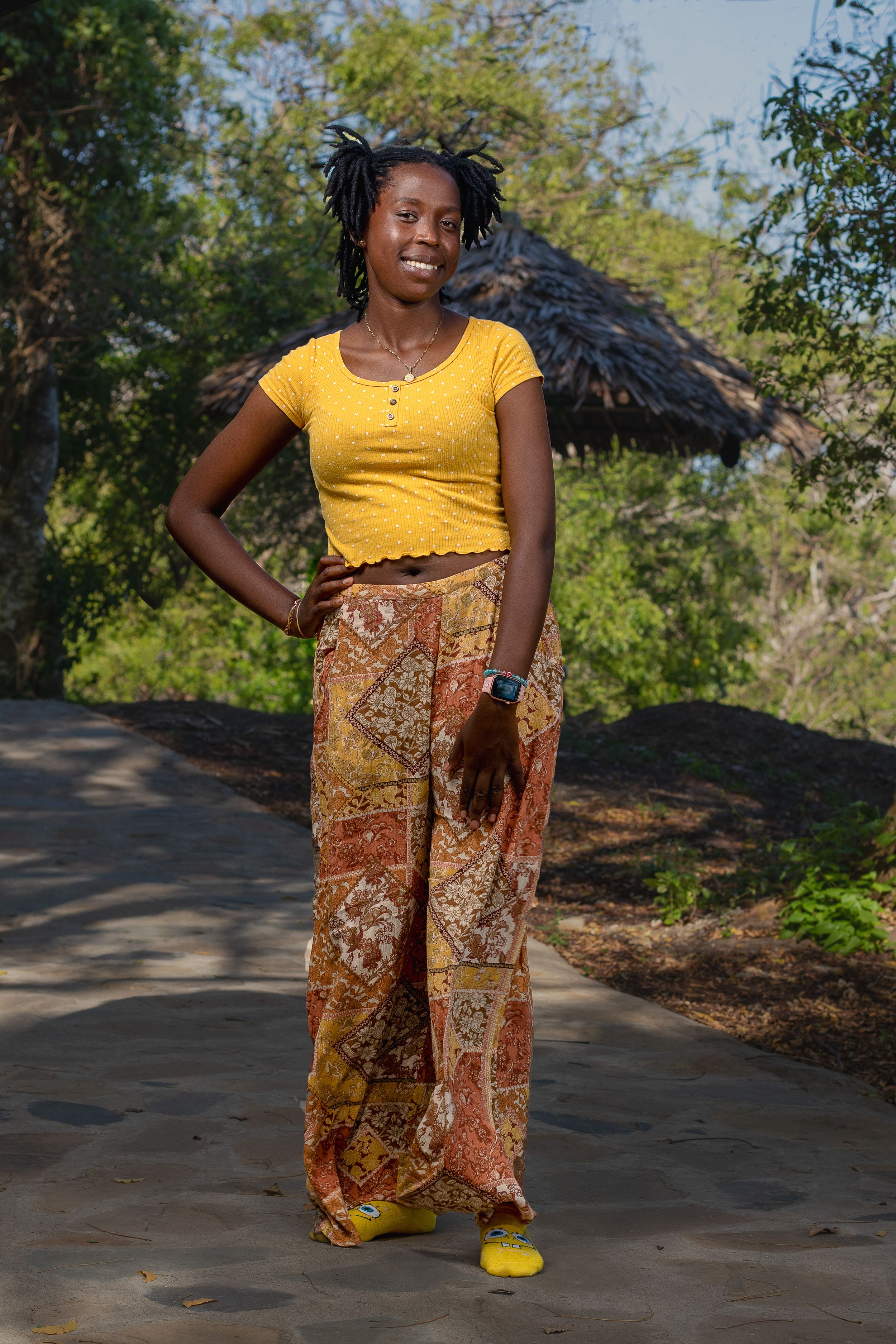 A happy lady smiling , stands  on a pavement in front of thatched hut