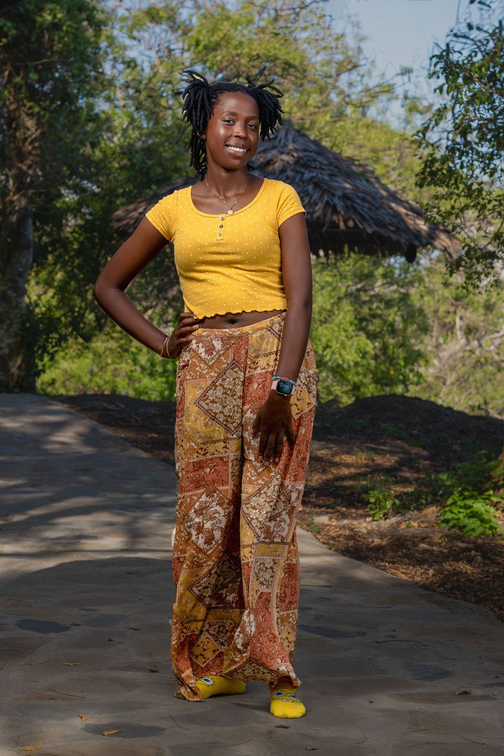 A happy lady smiling , stands  on a pavement in front of thatched hut