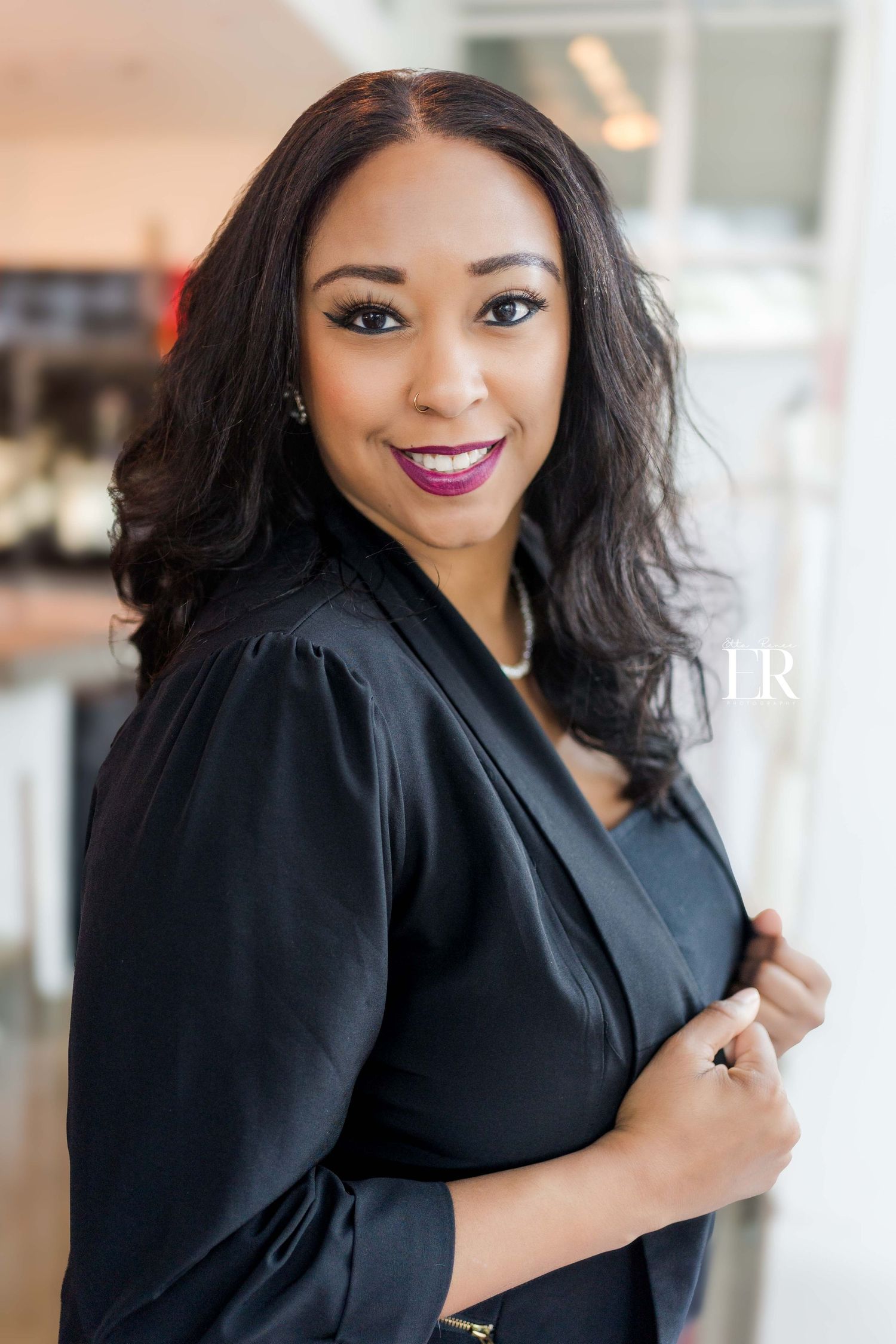 Portrait of a woman in black blazer smiling warmly in a bright indoor setting.