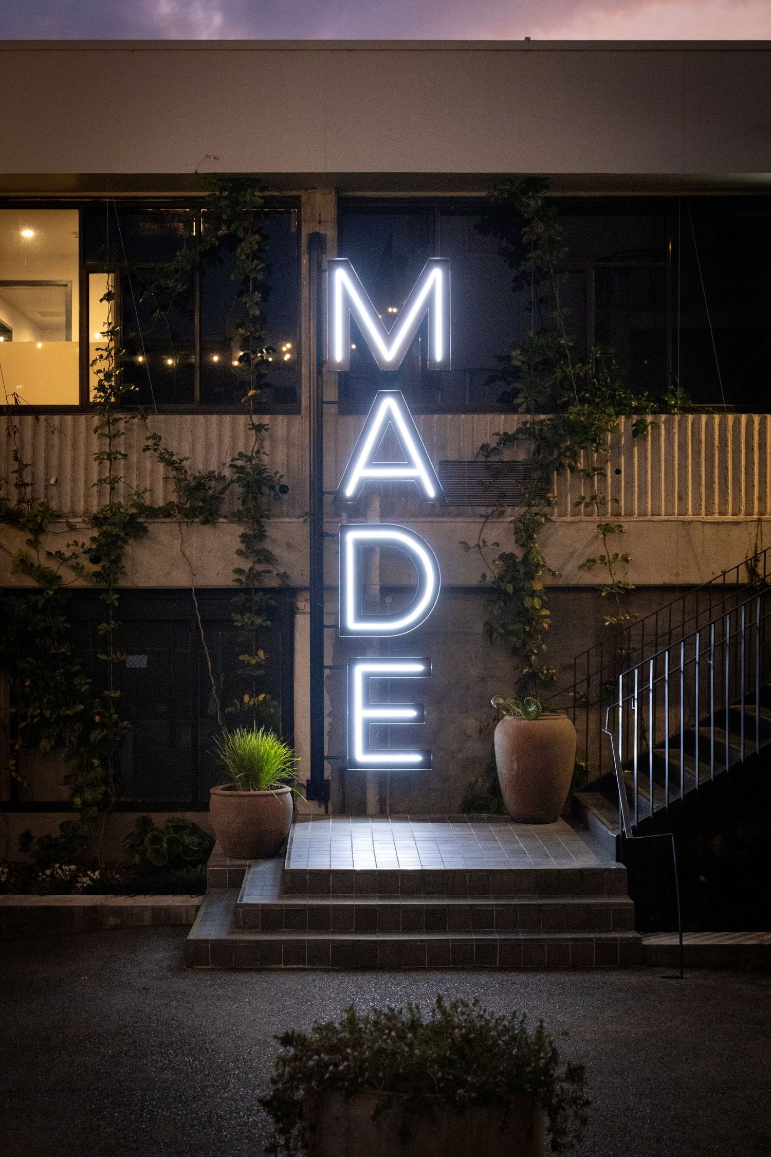 White neon sign reading “MADE” outside a stylish urban storefront at night, with stairs and potted plants nearby.