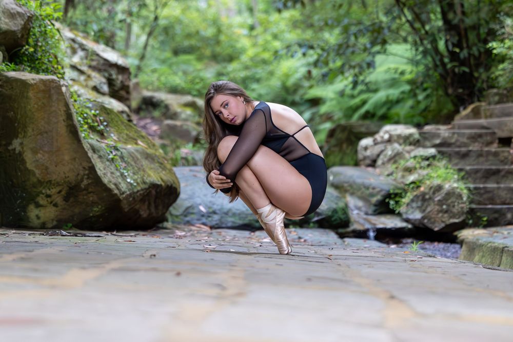 A person crouches in a graceful pose on a stone path surrounded by mossy rocks and lush greenery.