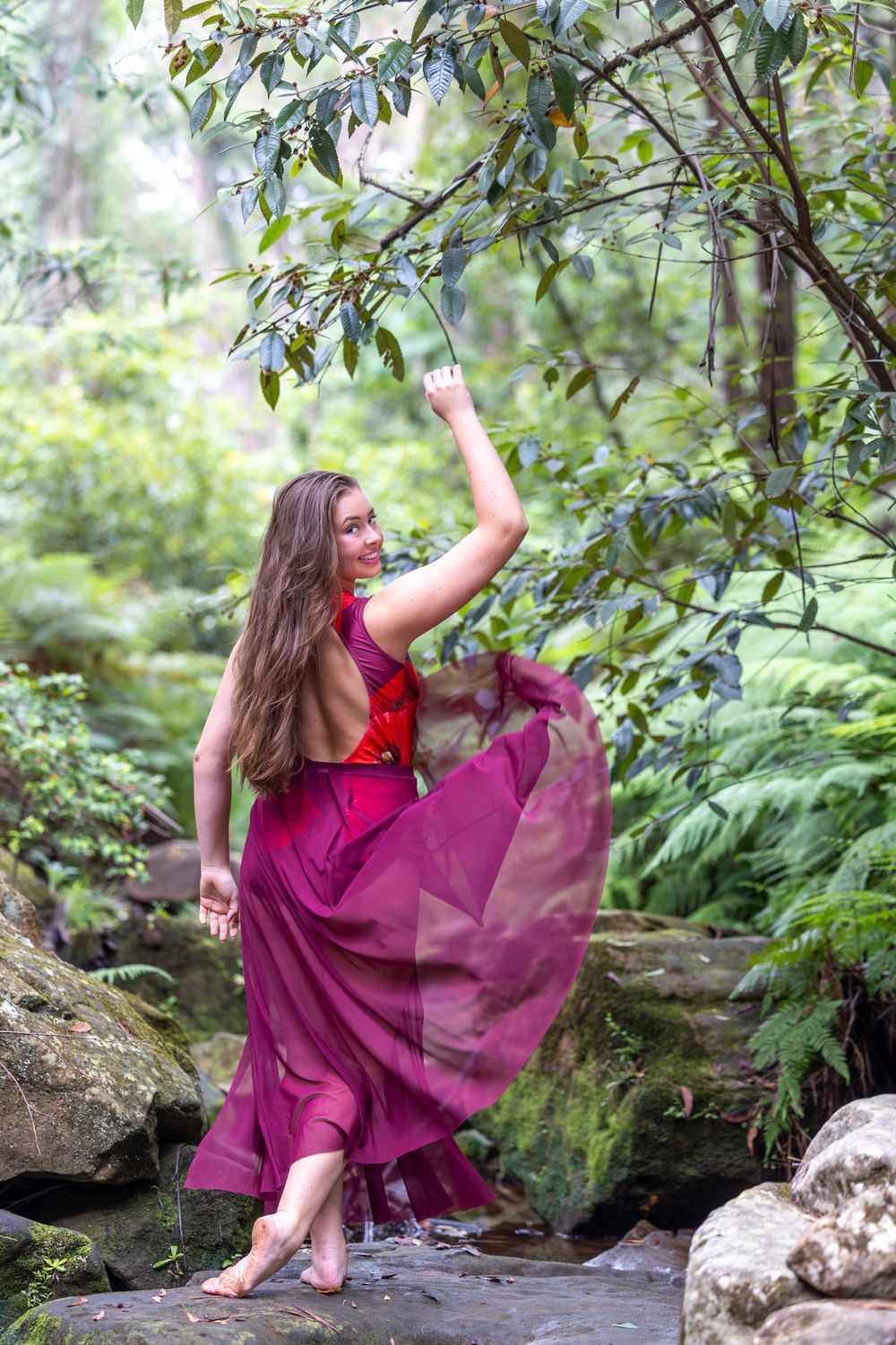 A person in a flowing magenta dress dances gracefully on mossy rocks in a lush forest setting.