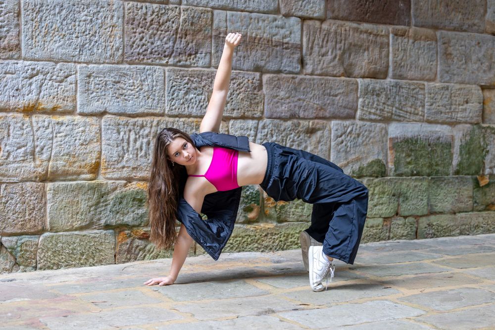 A dancer performs a dynamic breakdance move in front of a stone wall while wearing a pink top and dark pants.
