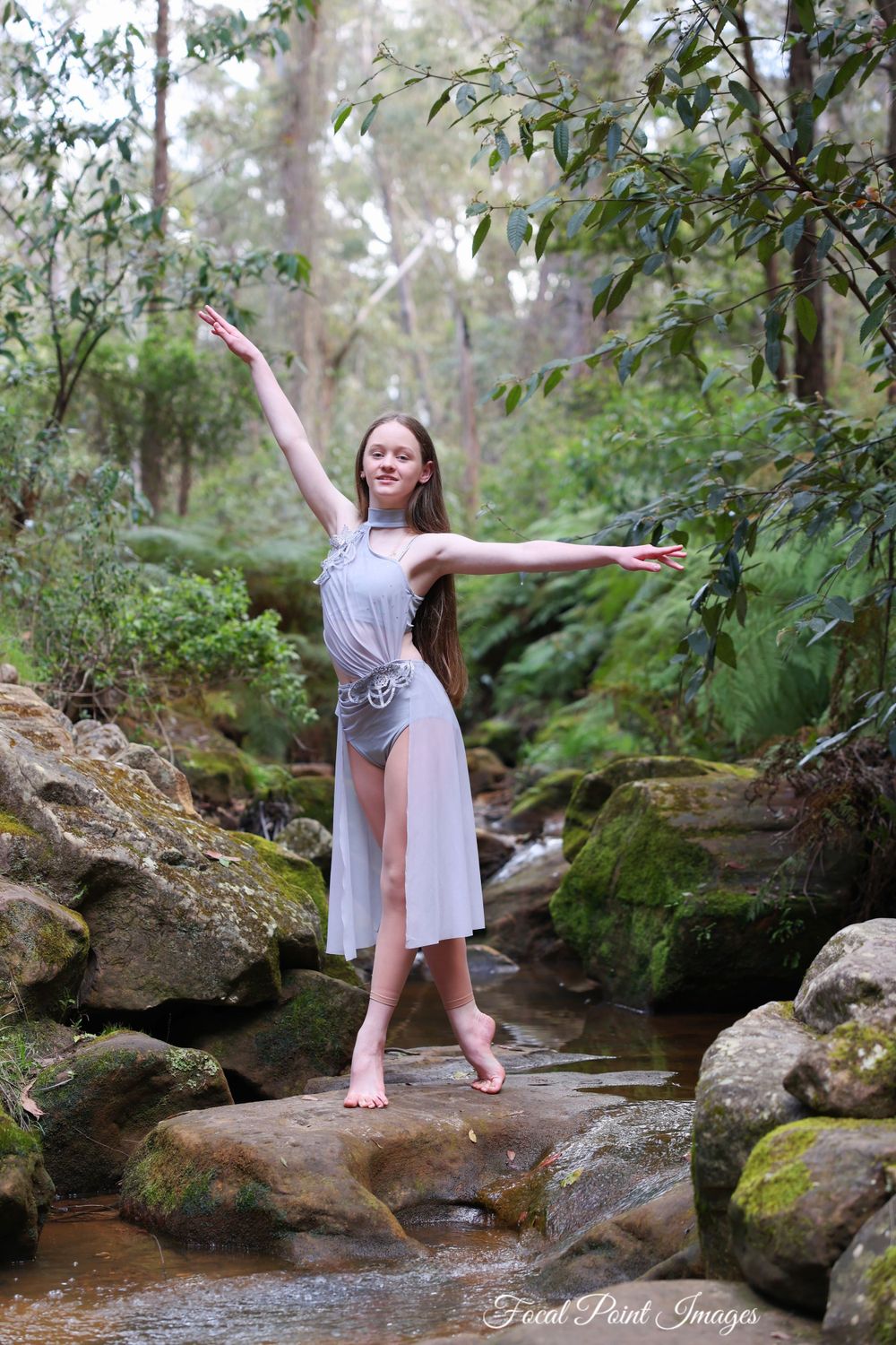 Barefoot dancer in flowing dress strikes elegant pose on mossy rocks in forest stream.